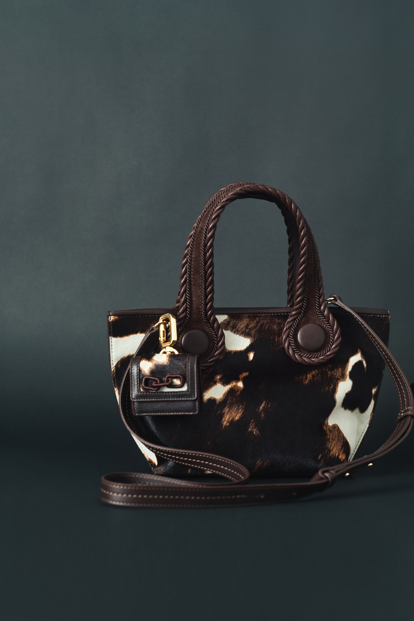 A dark brown and cream cow print pattern small tote bag with dark brown rope handles and strap  on a dark background. An airpods case in the same color and texture attached to the bag.