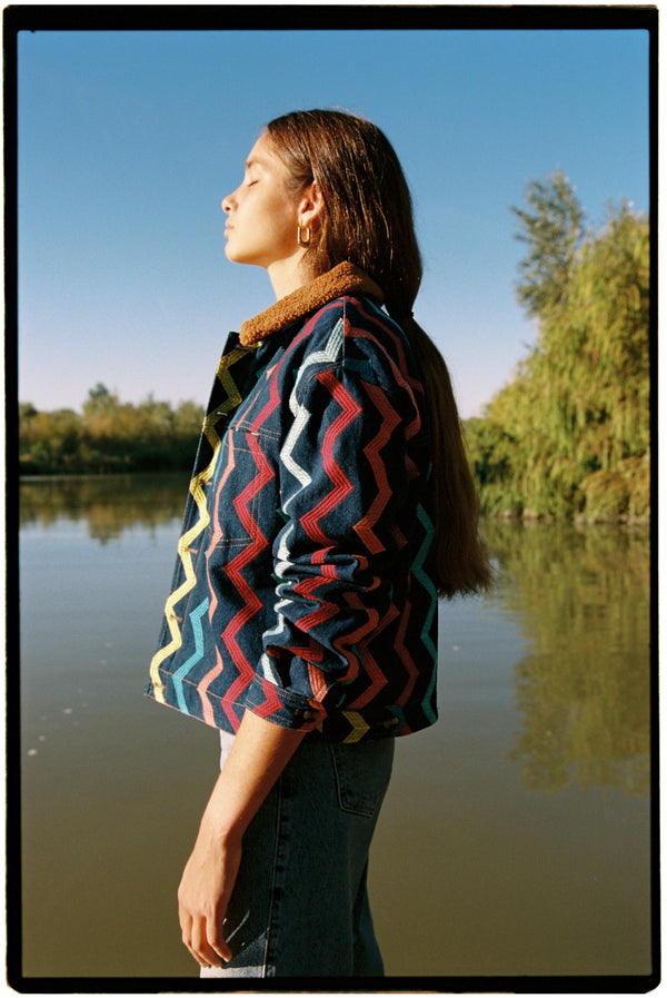 Michonet – Robe Denim Zig Zag, mannequin de profil portant une veste en jean colorée au bord de l'eau