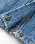 Detalle de primer plano de jeans de mezclilla azul desteñido, 100% algodón, con cremallera metálica y botones de remache expuestos, costuras en contraste naranja.