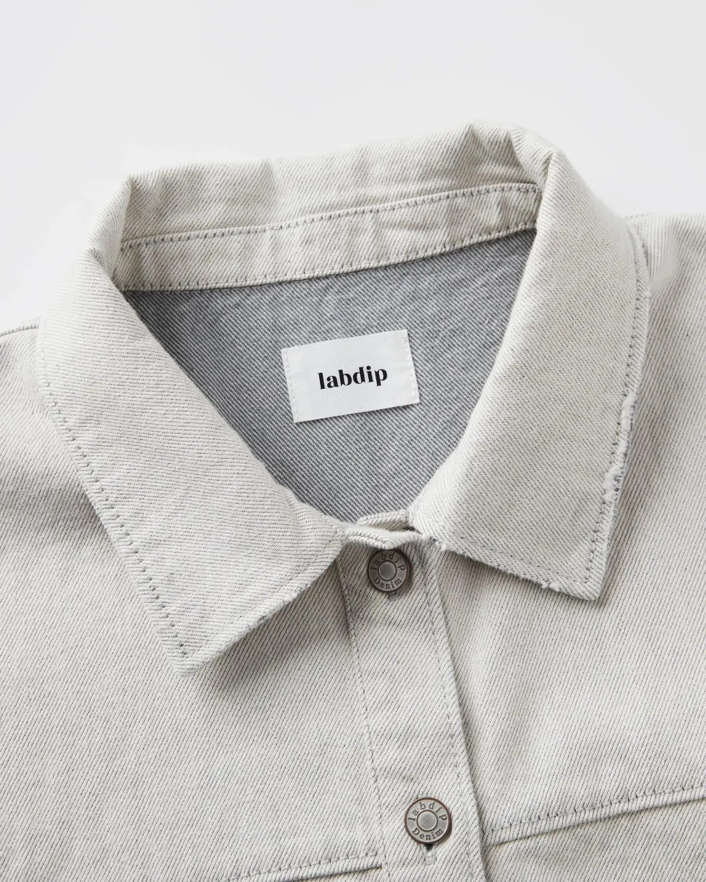 Light granite-colored undyed denim shirt collar with visible stitching and metal buttons, 100% cotton fabric with a structured and natural feel.