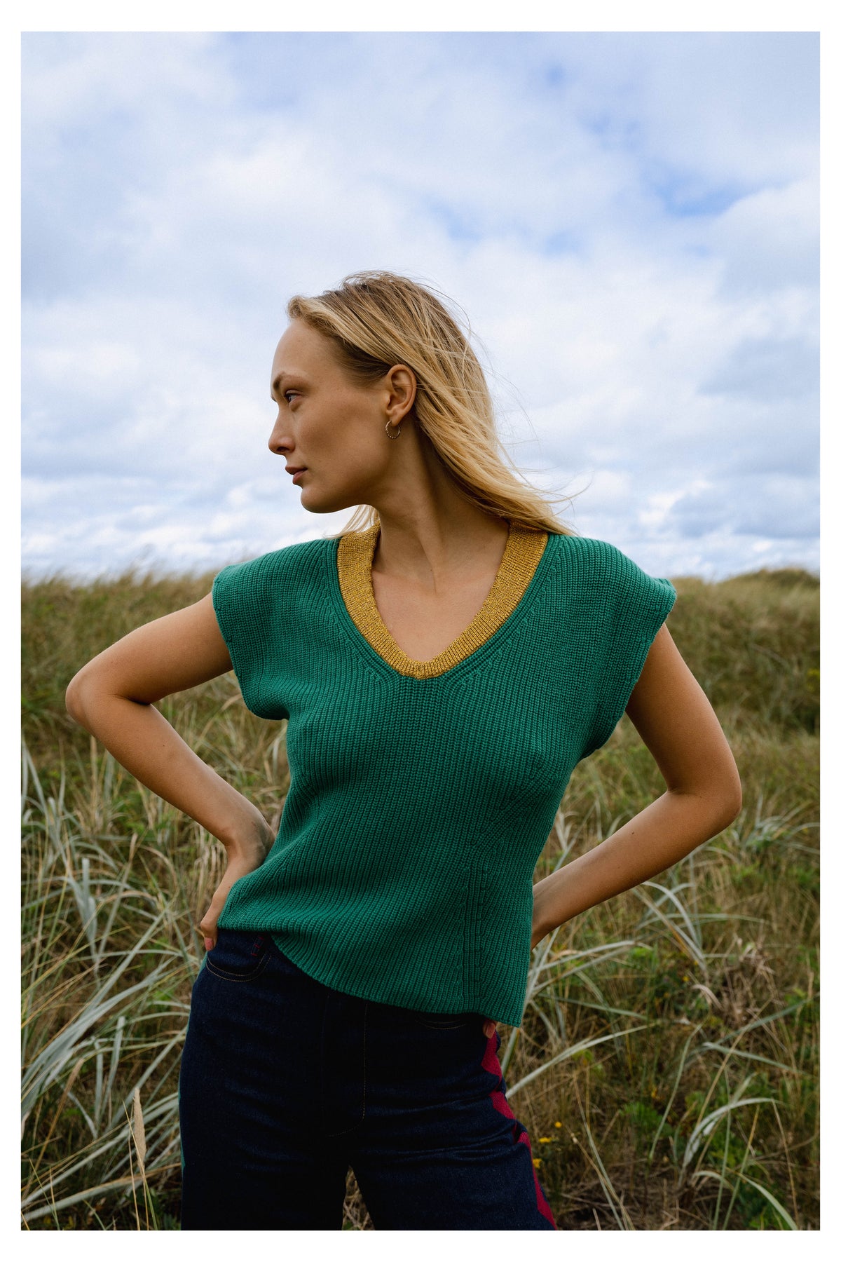 Michonet – Ärmellose Strickweste Knitted Green, Model in grünem Pullover und blauer Hose, im Profil im Gras posierend
