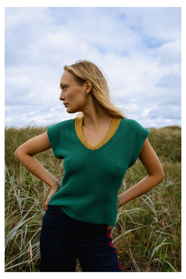 Michonet – Gilet Sans Manches Knitted Green, mannequin en pull vert et pantalon bleu, posant de profil dans l'herbe