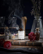 Suero L'Élixir du Marabout de Garancia colocado sobre un antiguo grimorio, rodeado de flores rojas, viales de boticario y un humo místico que evoca un laboratorio de brujería.