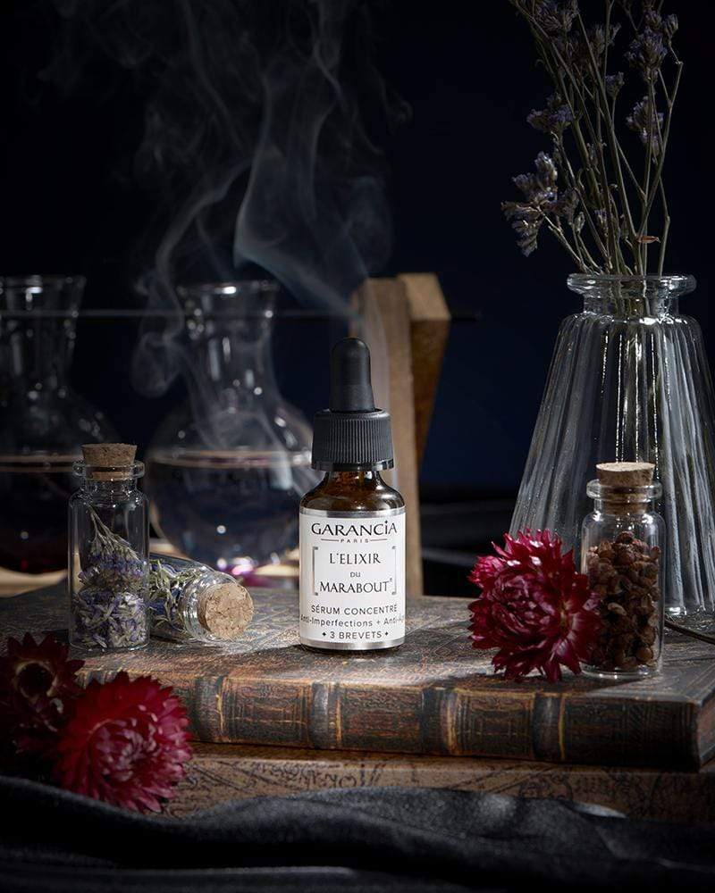 Suero L'Élixir du Marabout de Garancia colocado sobre un antiguo grimorio, rodeado de flores rojas, viales de boticario y un humo místico que evoca un laboratorio de brujería.