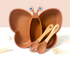 Plato infantil con forma de mariposa en color terracota con antenas y ojos, acompañado de cubiertos de madera grabados con "Alba", visto en primer plano.