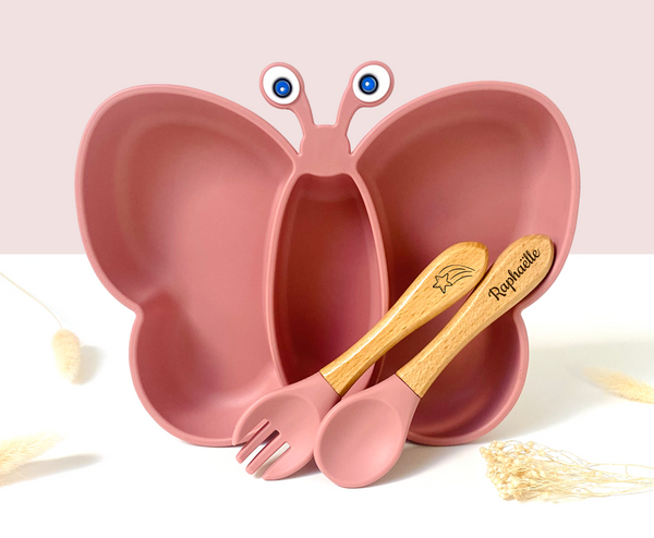 Léon l’assiette Papillon: set de vaisselle pour enfants rose en forme de papillon avec ustensiles bois gravés “Raphaëlle” posé dessus.