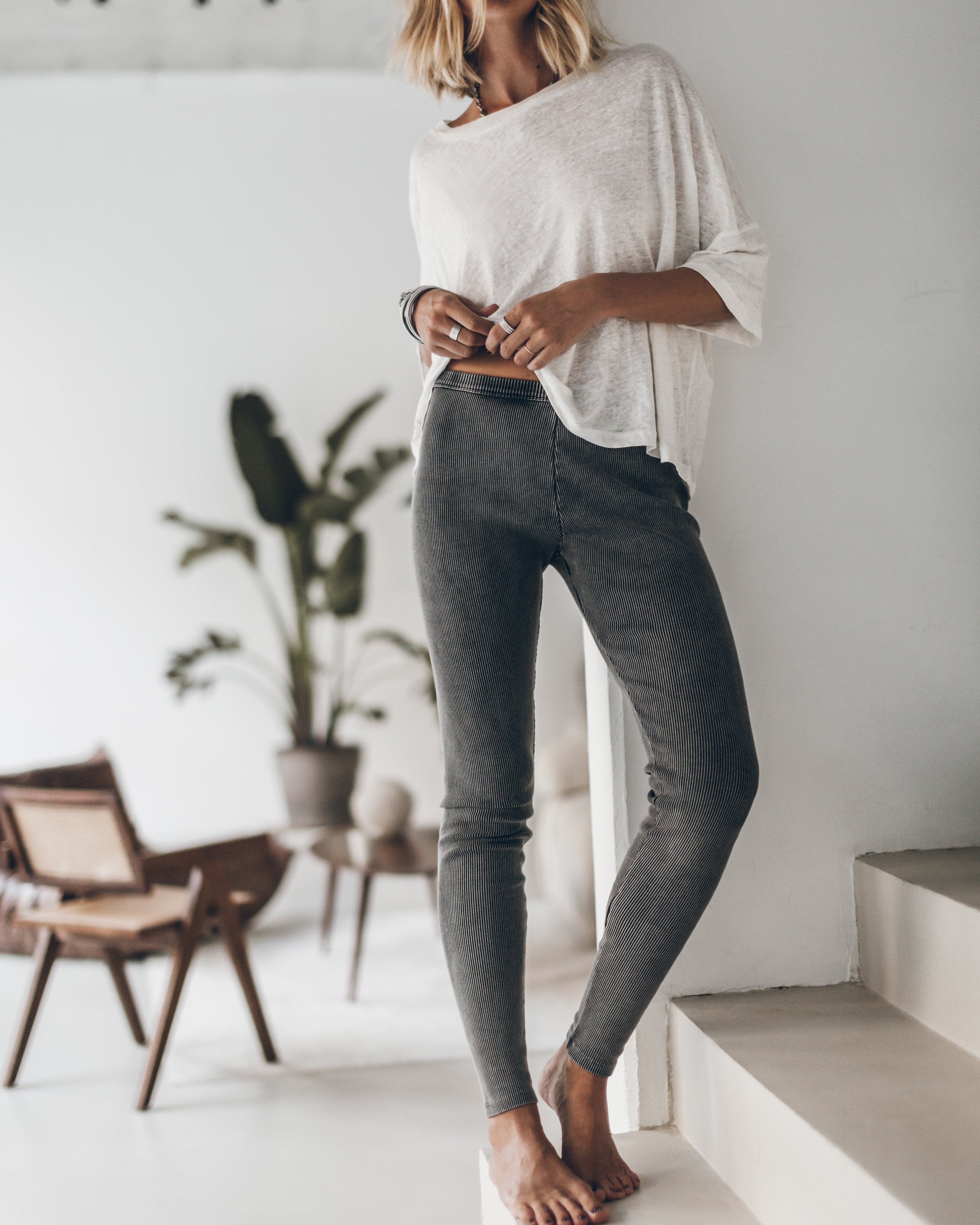 Person wearing a white oversized shirt and gray leggings standing on a staircase in a bright room with plants.